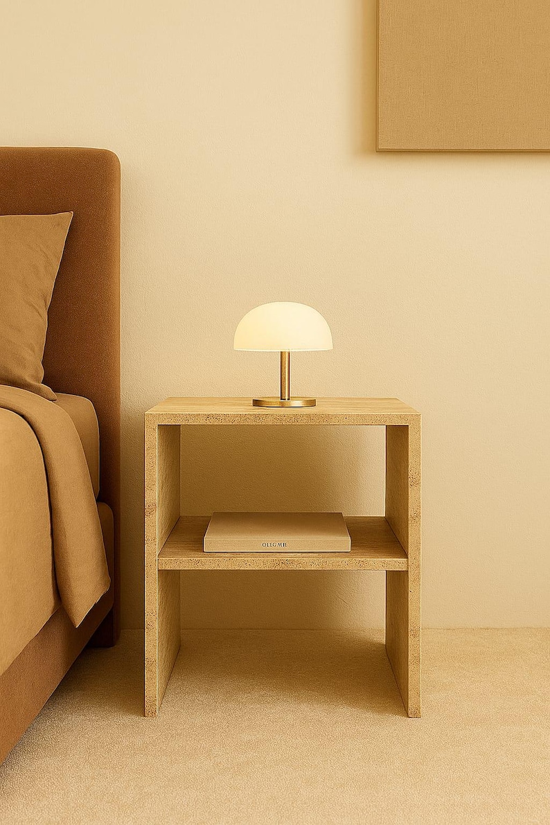 May include: A modern bedroom scene featuring a small, white, dome-shaped table lamp with a gold base on a light wood nightstand. A book is on the lower shelf. The bed and wall are a matching beige color.