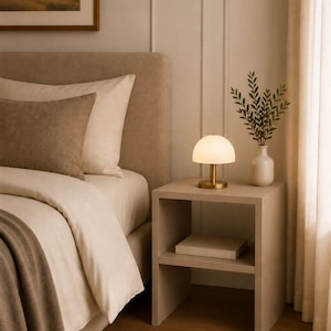 May include: A bedroom scene featuring a neutral color palette. A bedside table holds a lamp with a gold base and a white shade, a vase with greenery, and a book. The bed is made with beige linens and a brown throw.
