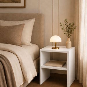 May include: A bedroom scene featuring a white nightstand with a small lamp and a book. A beige bed with pillows and a blanket is in the background. A framed landscape painting hangs on the wall, and a rug is on the floor.