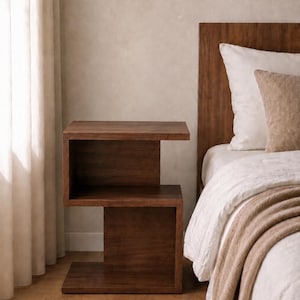 May include: A dark brown, modern nightstand with a unique, open design. The two-tiered side table is made of wood and has a minimalist aesthetic. The nightstand is placed next to a bed with white bedding and a brown headboard.
