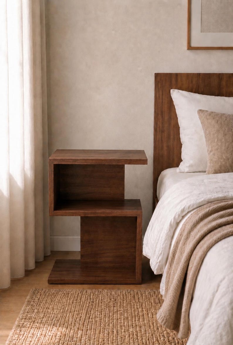 May include: A dark brown, modern nightstand with a unique, open design. The two-tiered side table is made of wood and has a minimalist aesthetic. The nightstand is placed next to a bed with white bedding and a brown headboard.