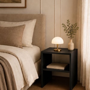 May include: A bedroom scene featuring a black nightstand with a small lamp and a vase of greenery. The bed has beige bedding and a brown throw blanket. A framed landscape painting hangs on the wall.