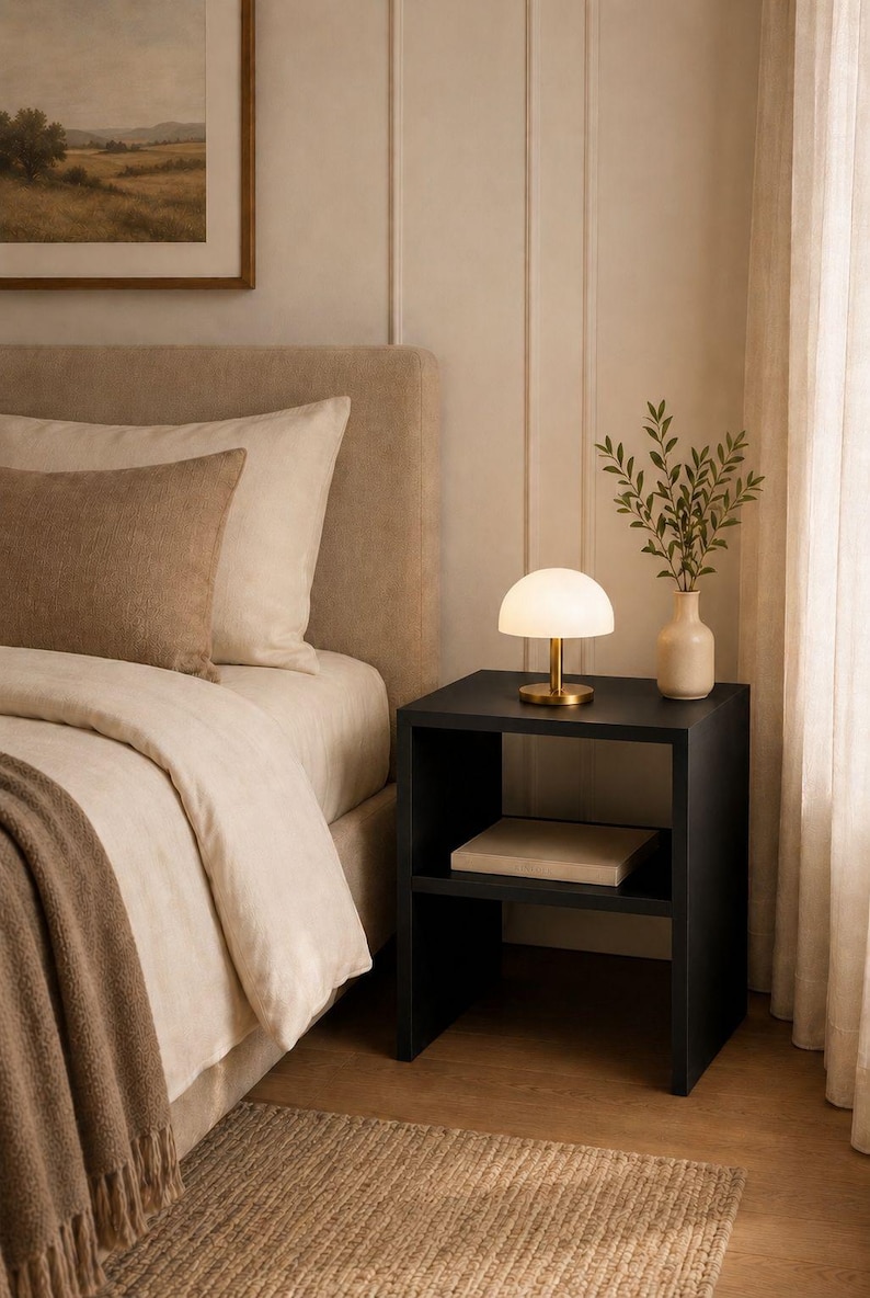 May include: A bedroom scene featuring a black nightstand with a small lamp and a vase of greenery. The bed has beige bedding and a brown throw blanket. A framed landscape painting hangs on the wall.