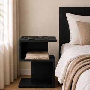 May include: A modern black nightstand with a unique, open design. The nightstand features two shelves, with a smartphone, glasses, and two books on display. The books are titled "The Kinfolk Home" and "Live Beautifully". The nightstand is next to a bed with white bedding.