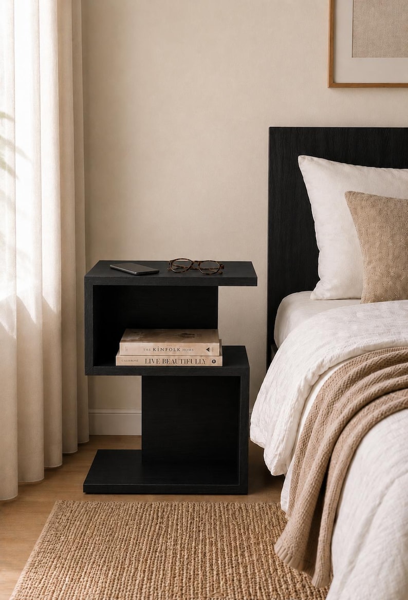 May include: A modern black nightstand with a unique, open design. The nightstand features two shelves, with a smartphone, glasses, and two books on display. The books are titled "The Kinfolk Home" and "Live Beautifully". The nightstand is next to a bed with white bedding.