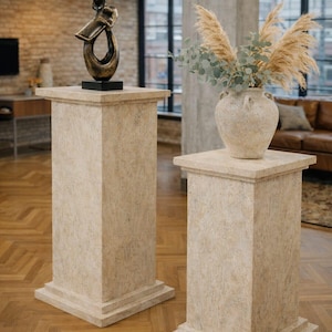 K&ouml;nnte beinhalten: Zwei beige Sockel mit quadratischen Oberseiten und Basen. Einer tr&auml;gt eine Bronzeskulptur, der andere eine Vase mit getrockneten Gr&auml;sern und Gr&uuml;n. Die Sockel stehen in einem Raum mit Parkettboden und gro&szlig;en Fenstern.