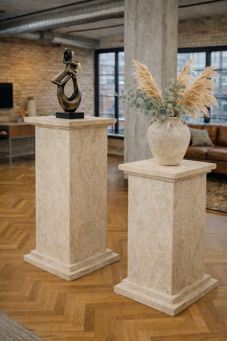 K&ouml;nnte beinhalten: Zwei beige Sockel mit quadratischen Oberseiten und Basen. Einer tr&auml;gt eine Bronzeskulptur, der andere eine Vase mit getrockneten Gr&auml;sern und Gr&uuml;n. Die Sockel stehen in einem Raum mit Parkettboden und gro&szlig;en Fenstern.
