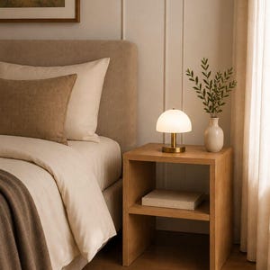 May include: A bedroom scene featuring a wooden nightstand with a lamp, a vase with greenery, and a book. The bed has a beige headboard, white bedding, and a brown throw. A framed landscape painting hangs on the wall.