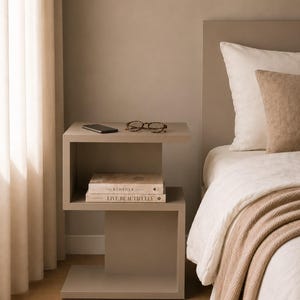 May include: A modern, taupe-colored nightstand with a geometric design. The nightstand holds a smartphone, eyeglasses, and two books titled "The Kinfolk Home" and "Live Beautifully." A bed with white linens and a beige throw is in the background.