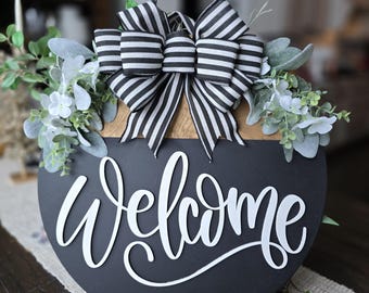 Op de afbeelding: Een ronde, zwarte houten plank met het woord "Welcome" in witte letters. Versierd met een zwart-wit gestreepte strik, groen en kleine witte bloemen. Het bord staat op een neutraal gekleurde tafelloper.
