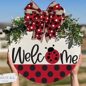 May include: A round wooden welcome sign with a red and black polka dot pattern. The sign features the word "Welcome" in black script with a ladybug in the center. A red and black polka dot bow with ladybugs is attached to the top of the sign.