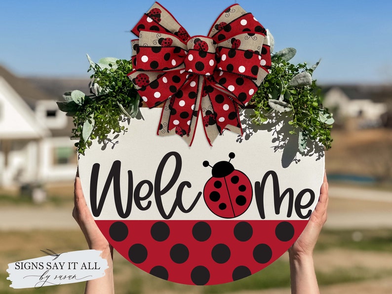 May include: A round wooden welcome sign with a red and black polka dot pattern. The sign features the word "Welcome" in black script with a ladybug in the center. A red and black polka dot bow with ladybugs is attached to the top of the sign.