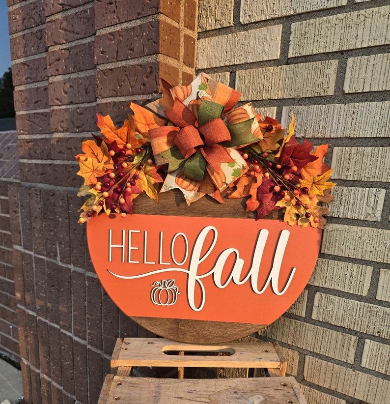 May include: A round wooden sign with a red background and white text that says 'hello fall'. The sign is decorated with a fall-themed wreath made of orange, red, and yellow leaves, berries, and a green and orange bow. A small pumpkin is cut out of the sign below the text.