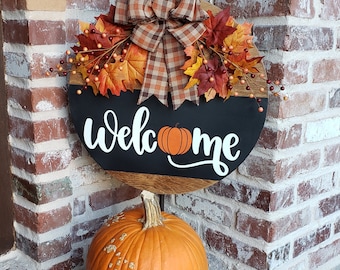 Puede incluir: Un letrero de puerta redondo de madera con una sección pintada de negro que incluye el texto blanco welcome y un gráfico de calabaza naranja. El letrero está decorado con un lazo a cuadros marrón y hojas y bayas de otoño artificiales. Una calabaza naranja grande se sitúa debajo.