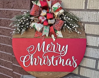 May include: A round wooden door hanger with a red background and the words "Merry Christmas" in white. The hanger is decorated with a large red, gold, and green bow with white glitter.