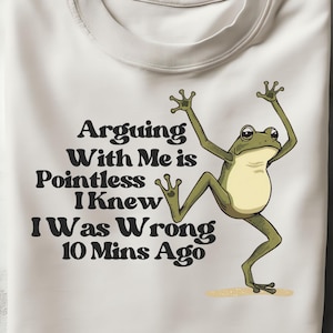May include: A folded, off-white t-shirt with the text "Arguing With Me is Pointless I Knew I Was Wrong 10 Mins Ago" and a cartoon frog illustration in green and tan.