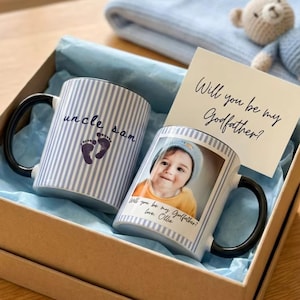 Puede incluir: Una caja de regalo con dos tazas, una tarjeta, una manta y un osito de peluche. Las tazas tienen asas negras y están decoradas con rayas azules y blancas. Una taza tiene una foto de un bebé. La tarjeta dice: "¿Quieres ser mi padrino?"
