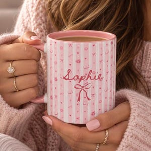 May include: A ceramic mug with a pink and white vertical striped pattern featuring small icons of bows, books, and glasses. The name Sophie is printed in a cursive pink script above a larger pink bow graphic on the front of the mug.