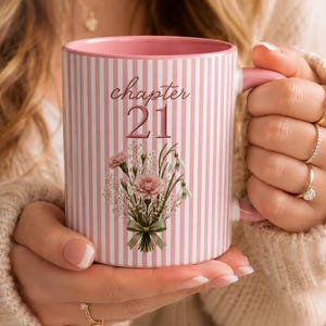 May include: A ceramic mug with a pink and white vertical striped pattern. The front features the text 'chapter 21' in a script font above a floral bouquet illustration. The mug has a solid pink interior and a pink handle.