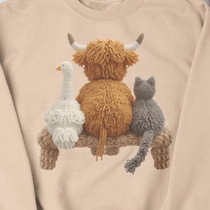 Könnte beinhalten: Beiges Sweatshirt mit Strickdesign einer Gans, einer Hochlandkuh und einer Katze auf einer Bank. Die Kuh ist braun, die Gans ist weiß und die Katze ist grau. Das Sweatshirt ist von Winteraccessoires umgeben.
