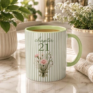 May include: A ceramic mug with a light green and white vertical striped pattern. The front features the text "chapter 21" in a dark green script font above a floral illustration of pink carnations and white flowers tied with a green ribbon.