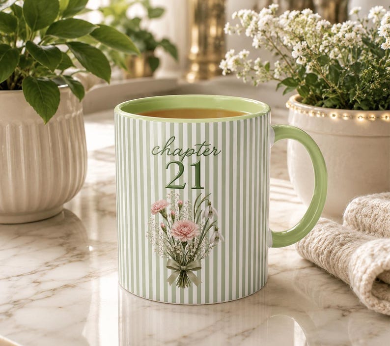 May include: A ceramic mug with a light green and white vertical striped pattern. The front features the text "chapter 21" in a dark green script font above a floral illustration of pink carnations and white flowers tied with a green ribbon.