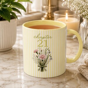 May include: A ceramic mug with a pale yellow and white vertical striped pattern. The front features the text "chapter 21" in a cursive font above a bouquet of pink carnations and white flowers tied with a sage green ribbon. The mug has a yellow handle.