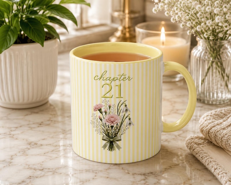 May include: A ceramic mug with a pale yellow and white vertical striped pattern. The front features the text "chapter 21" in a cursive font above a bouquet of pink carnations and white flowers tied with a sage green ribbon. The mug has a yellow handle.