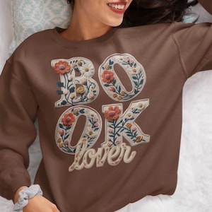 Może przedstawiać: Brązowa bluza z napisem "BOOK LOVER" w kwiatowym wzorze. Litery są białe z pomarańczowymi i żółtymi kwiatami oraz zielonymi liśćmi. Bluza jest w jednolitym brązowym kolorze.