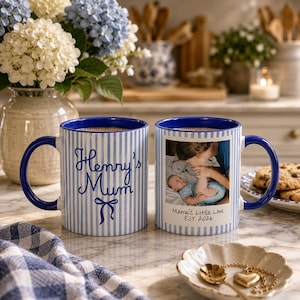 Peut inclure: Deux mugs en c&eacute;ramique blanche &agrave; rayures et anses bleues. L'un porte l'inscription "Henry's Mum" et un n&oelig;ud. L'autre mug a une photo et le texte "Mama's Little Love EST 2026".