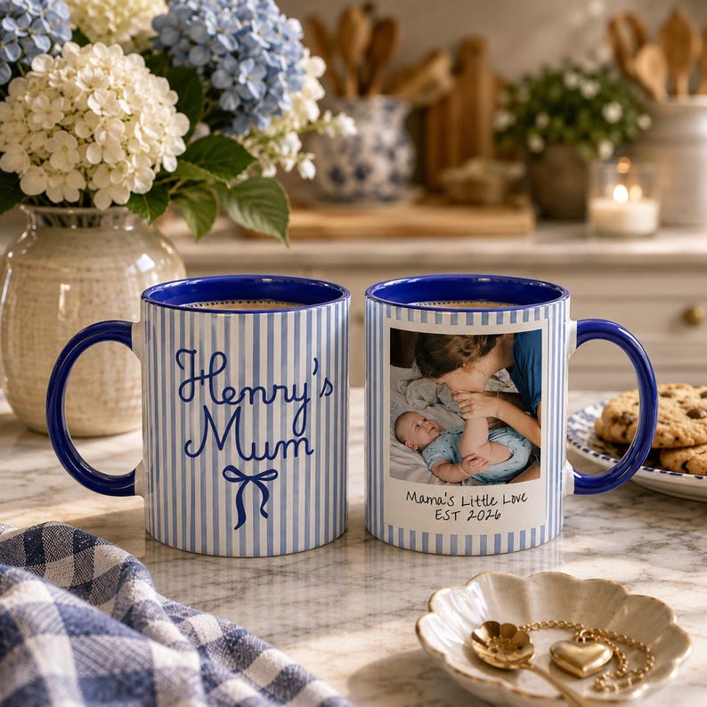 Peut inclure: Deux mugs en c&eacute;ramique blanche &agrave; rayures et anses bleues. L'un porte l'inscription "Henry's Mum" et un n&oelig;ud. L'autre mug a une photo et le texte "Mama's Little Love EST 2026".