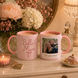 Peut inclure: Deux mugs &agrave; rayures roses et blanches avec des anses roses. L'un des mugs porte l'inscription "Bella's Mum" et un n&oelig;ud. L'autre mug porte une photo et l'inscription "love you mama". Les deux mugs sont remplis de caf&eacute;.