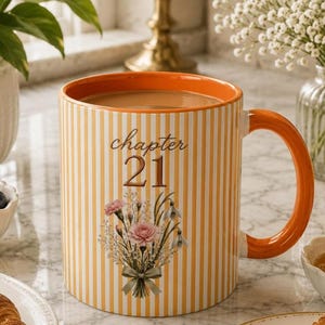 May include: A ceramic mug with a white and yellow vertical striped pattern, featuring the text "chapter 21" in brown script and block lettering above a floral bouquet illustration of pink carnations and white flowers tied with a green ribbon.