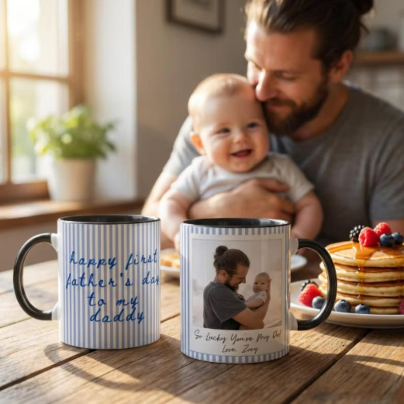 May include: Two white ceramic mugs with black handles and rims. One mug has blue vertical stripes and the text "Happy First Father's Day to my Daddy". The other mug has a photo and the text "So Lucky You're My Dad Love, Zoey".