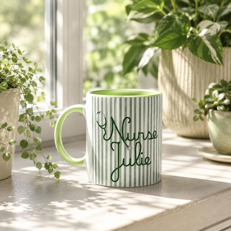 May include: A white and green striped ceramic mug with the text "Nurse Julie" and a stethoscope graphic. The mug has a green handle and interior. The mug is sitting on a white surface near a window with plants.