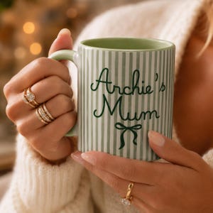 Peut inclure: Une tasse en c&eacute;ramique &agrave; rayures vertes et blanches avec l'inscription "Archie's Mum" en &eacute;criture vert fonc&eacute;. La tasse a une anse et un int&eacute;rieur verts. Un motif de n&oelig;ud est en dessous du texte. La tasse est tenue par une personne portant des bagues.