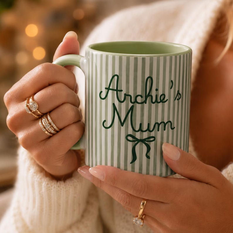 Peut inclure: Une tasse en c&eacute;ramique &agrave; rayures vertes et blanches avec l'inscription "Archie's Mum" en &eacute;criture vert fonc&eacute;. La tasse a une anse et un int&eacute;rieur verts. Un motif de n&oelig;ud est en dessous du texte. La tasse est tenue par une personne portant des bagues.