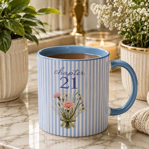 May include: A light blue and white striped ceramic mug with a blue handle and rim. The front features the text "chapter 21" in blue script and block lettering above a floral bouquet illustration containing pink carnations and white baby's breath.