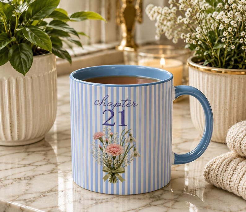 May include: A light blue and white striped ceramic mug with a blue handle and rim. The front features the text "chapter 21" in blue script and block lettering above a floral bouquet illustration containing pink carnations and white baby's breath.