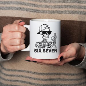 May include: White ceramic mug featuring a black and white illustration of a skeleton wearing sunglasses and a baseball cap. The text "SIX SEVEN" is printed below the skeleton. The mug has a white handle.