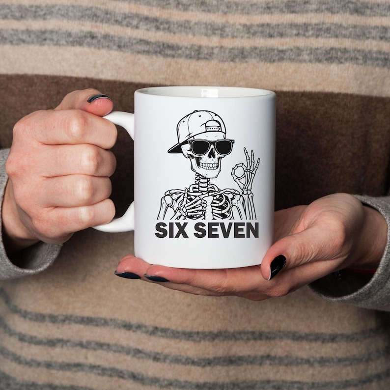 May include: White ceramic mug featuring a black and white illustration of a skeleton wearing sunglasses and a baseball cap. The text "SIX SEVEN" is printed below the skeleton. The mug has a white handle.