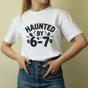 May include: White t-shirt with the phrase "HAUNTED BY 6-7" in black, featuring ghost and bat graphics. The shirt is a short-sleeved crew neck style. The text and graphics are centered on the front of the shirt.