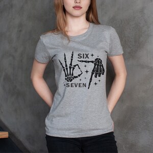 May include: A gray t-shirt featuring a black graphic of skeleton hands forming the numbers six and seven, with the words "SIX" and "SEVEN" above and below the hands. The shirt is a crew neck style.
