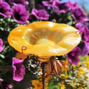 May include: A yellow glass bird feeder with copper wire accents and a decorative metal base. The feeder is shaped like a flower and is surrounded by purple and yellow flowers.