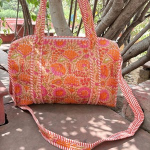 Könnte beinhalten: Eine gesteppte Reisetasche mit einem lebendigen Blumenmuster in Orange- und Rosatönen auf hellem Grund. Die Tasche verfügt über zwei lange Schulterriemen und einen Reißverschluss, die mit einem weiß-orange gestreiften Stoffbesatz verziert sind.