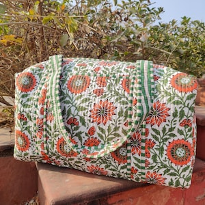 Könnte beinhalten: Eine weiße, gesteppte Tragetasche mit einem orangefarbenen und grünen Blumenmuster. Die Tasche hat grün gestreifte Griffe und Verzierungen. Die rechteckige Tasche scheint aus Baumwolle oder einem ähnlichen Stoff zu sein.