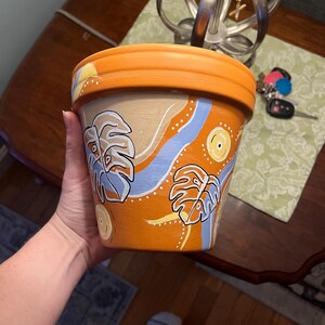 May include: Hand-painted orange ceramic flower pot with a wide rim. The pot features a beige background with blue and yellow accents, and stylized leaf designs. The pot is designed for indoor or outdoor use.