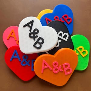 May include: A collection of heart-shaped decorations in various colors, including red, orange, green, blue, and black. Each heart features the letters "A&B" in contrasting colors, creating a personalized and festive design.