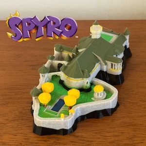 Può includere: Modello stampato in 3D di un castello della serie di videogiochi Spyro. Il modello presenta un tetto verde, alberi gialli e una piscina blu. Il logo Spyro è visibile nell'angolo in alto a sinistra.
