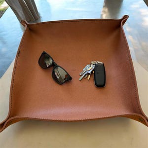 May include: A square tan leather valet tray with raised corners secured by metal fasteners. Inside the tray, there is a pair of black-framed sunglasses with dark lenses and a set of metal keys attached to a black plastic key fob.